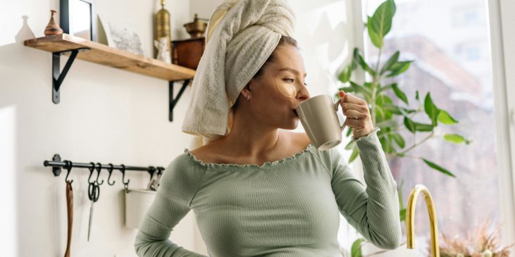 Schwangere mit Handtuch auf dem Kopf steht in der Küche und trinkt aus einer Tasse