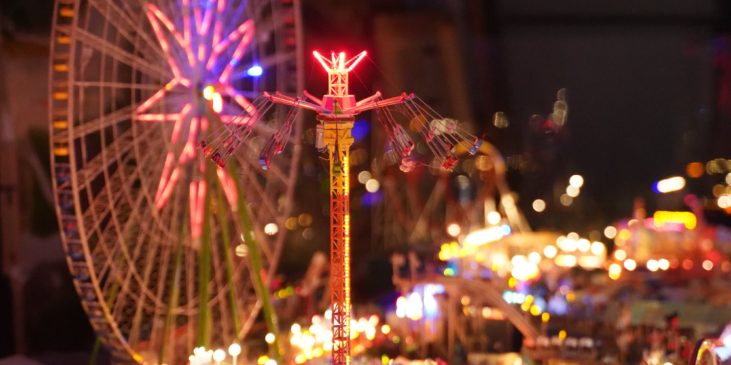 Die Wiesn bei Nachtstimmung, bunte Lichter erscheinen von den Fahrgeschäften und hellen die Atmosphäre auf. Im Vordergrund sieht man einen freien Fall Tower (Fahrgeschäft).