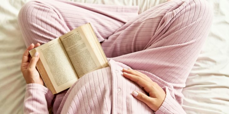 Schwangere sitzt, mit Buch in der Hand, im Bett und hält ihren Bauch