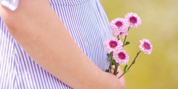 Der Bildausschnitt zeigt den schwangeren Bauch einer Frau, diese hällt ihn sanft in ihren Händen und in der Hand fünf kleine Rosa-Lila Blumen, der Hintergrund ist verschwommen, man erkennt dennoch eine grüne Wiese