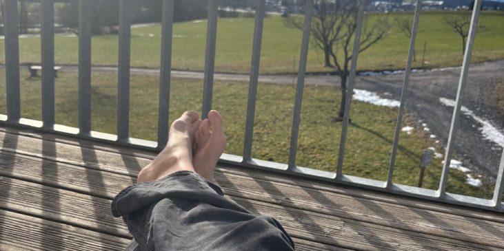 ausgestreckte Füße auf dem Balkon, im Hintergrund sieht man eine Wiese und Berge