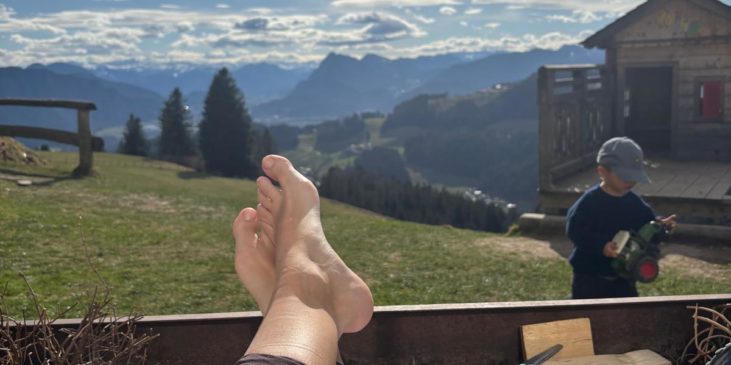 Füße lehnen auf einem Hochbeet, im Hintergrund spielt ein Kind und man sieht Berge und Wolken