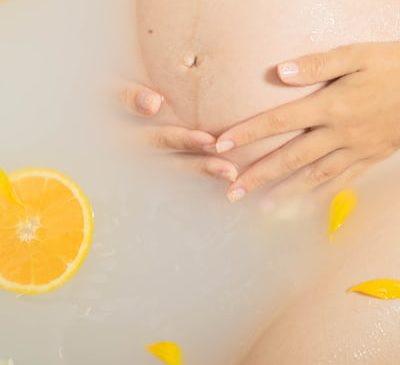 Eine schwangere Frau kurz vor der Wassergeburt in der Geburtswanne, man sieht den Bauch, ihre Hände und Orangen im Wasser