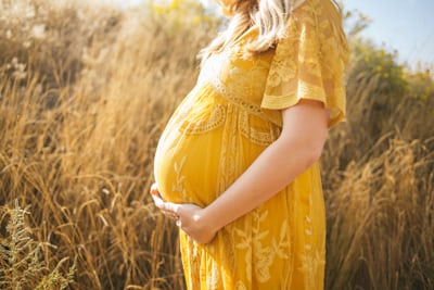 Ein schwangere Frau steht im gelben Sommerkleid in der Natur und haelt sich ihren BabyBauch