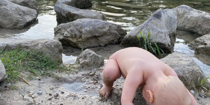 nacktes Kleinkind sitzt geduckt am Ufer eines Sees und schaut durch seine Beine hindurch