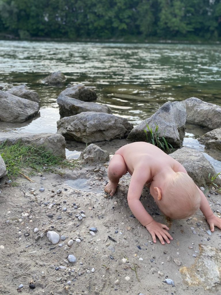 nacktes Kleinkind sitzt geduckt am Ufer eines Sees und schaut durch seine Beine hindurch