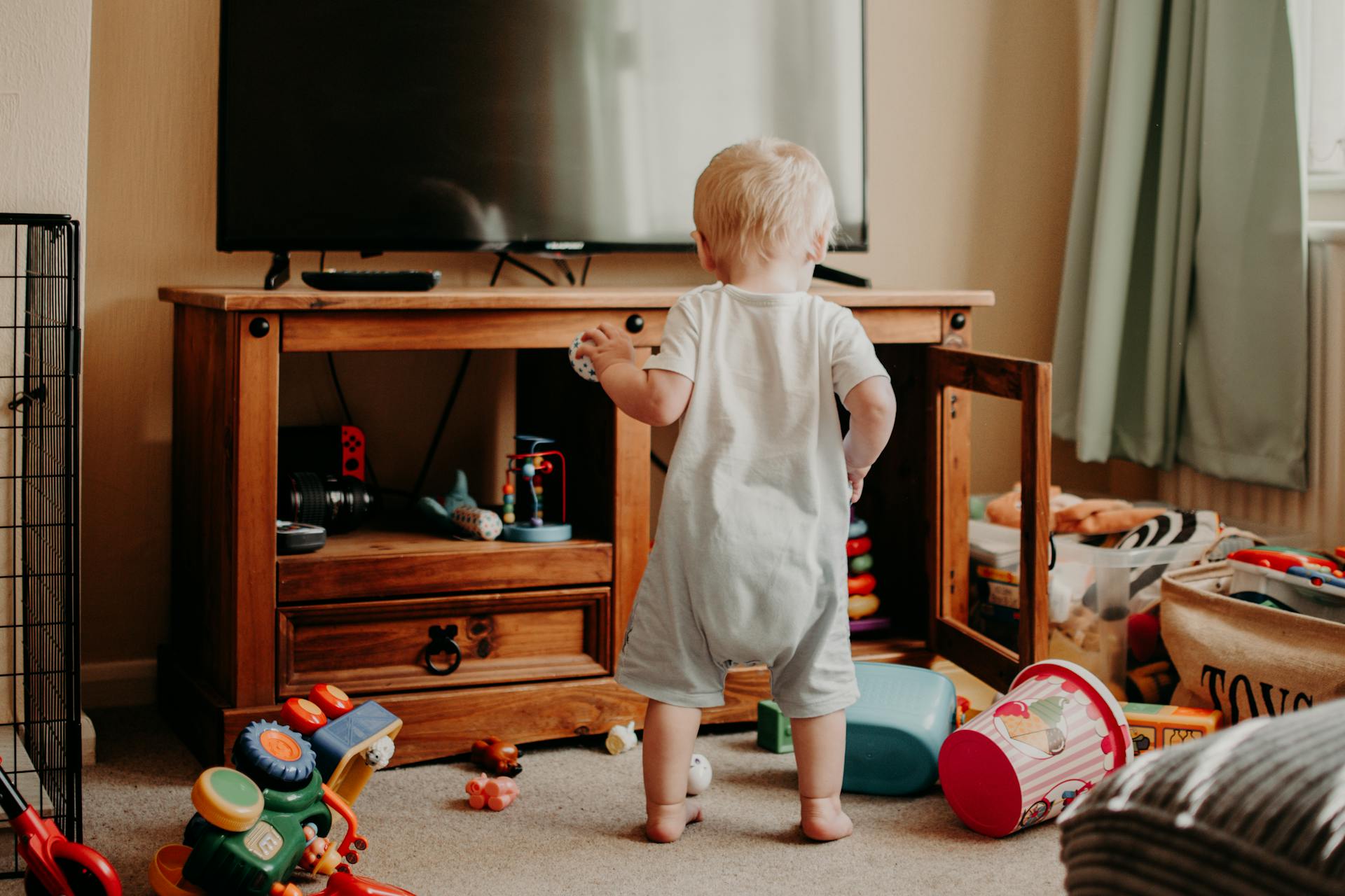 Kleinkind steht im Wohnzimmer zwischen verstreutem Spielzeug – typischer Alltag mit Baby und Spielchaos