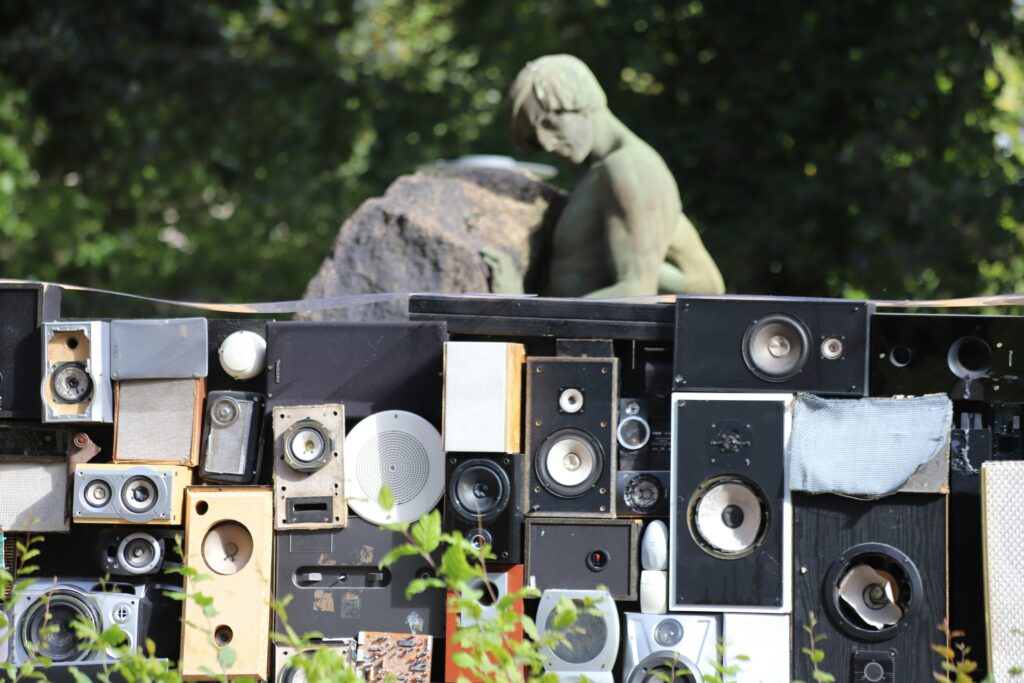 Man sieht mehrere Musikboxen, draußen, mit Natur und einer Steinstatue im Hintergrund
