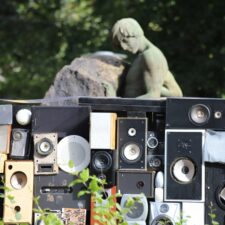 Man sieht mehrere Musikboxen, draußen, mit Natur und einer Steinstatue im Hintergrund