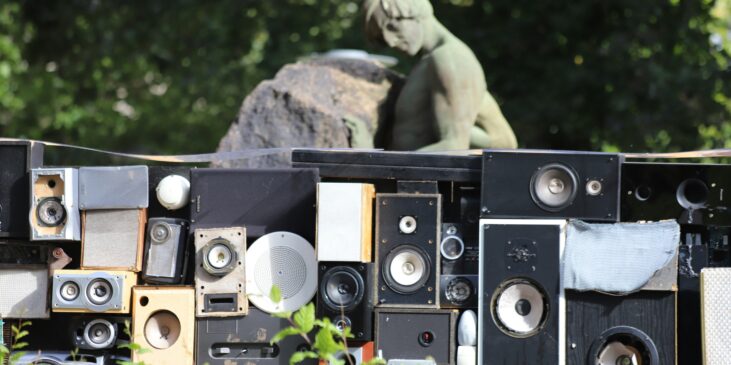 Man sieht mehrere Musikboxen, draußen, mit Natur und einer Steinstatue im Hintergrund