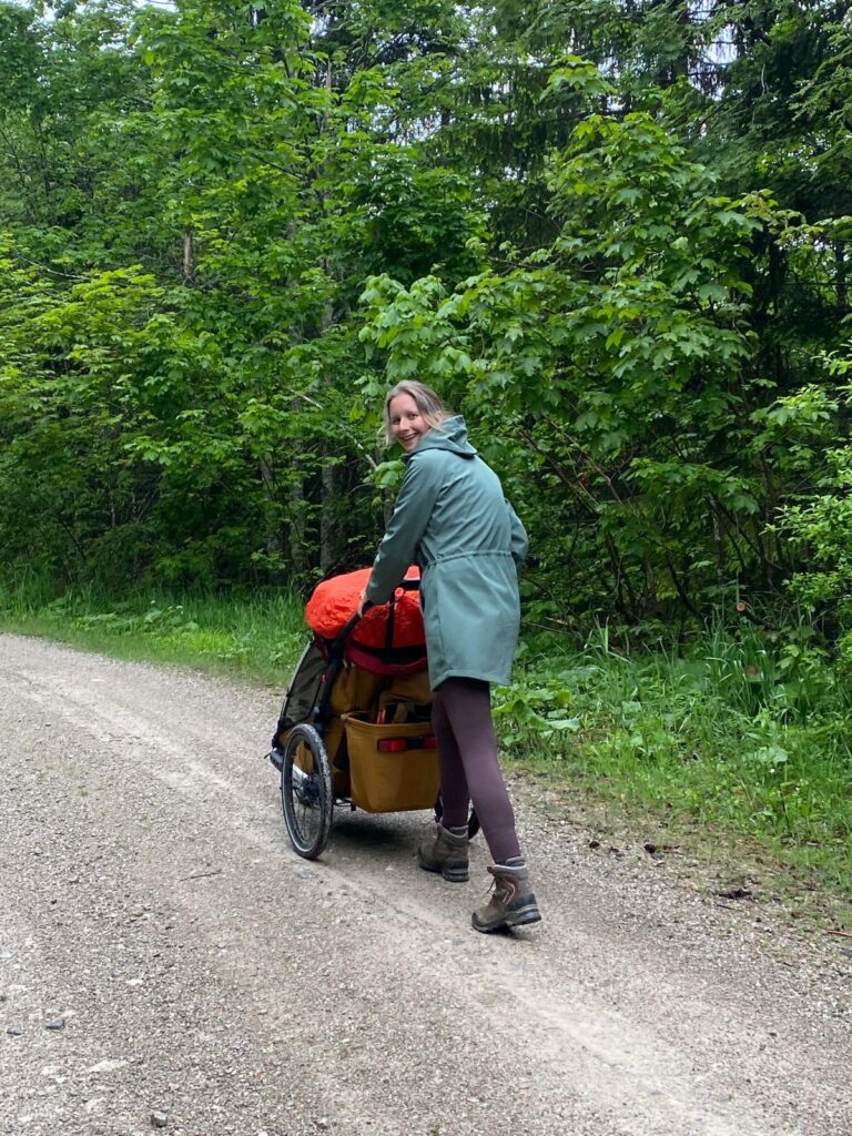 Frau mit Kinderwagen spaziert auf einem Waldweg und dreht sich zur Kamera