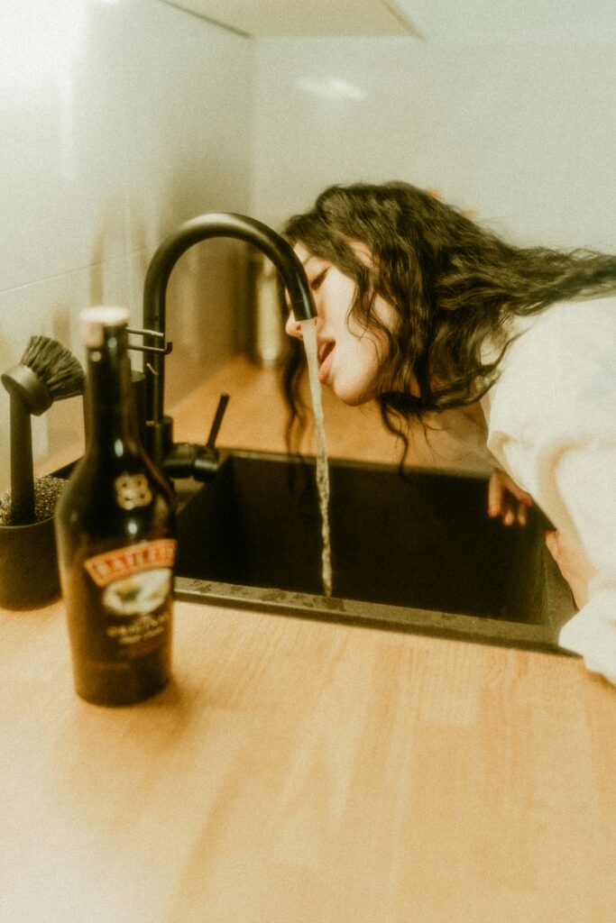 Frau, die Wasser aus einem Wasserhahn trinkt, daneben eine unberührte Flasche Alkohol