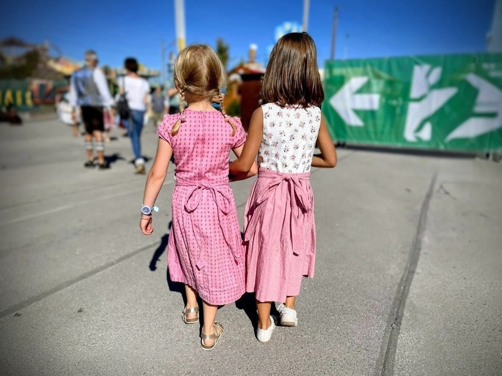 Zwei junge Mädchen in rosa Dirndln gehen Hand in Hand über einen Kirmesplatz, und zeigen ihre enge Freundschaft.